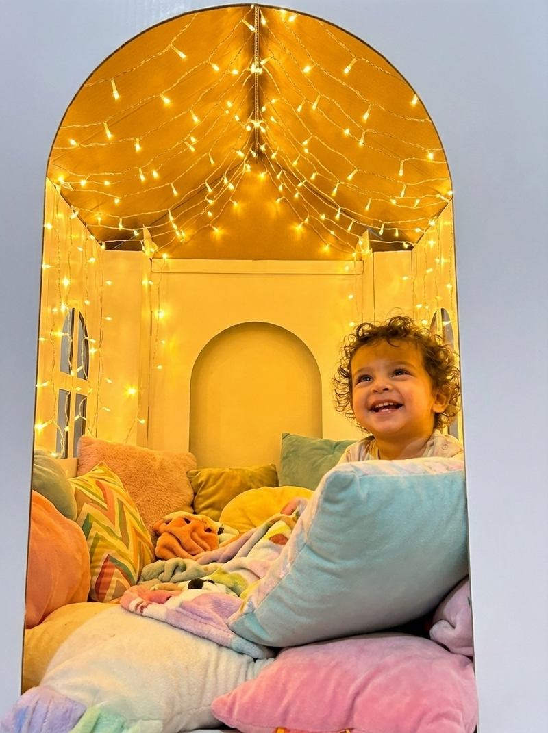Child sitting on a bed with colorful pillows and blankets, surrounded by fairy lights.