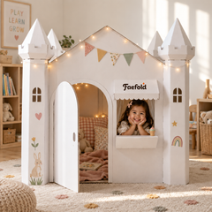 A child's white castle playhouse with decorative bunting, fairy lights, and a small window seat, shown in a warm, decorated children's room. A young girl is smiling from inside the playhouse, and the name 'Faefold' is printed on the front awning.
