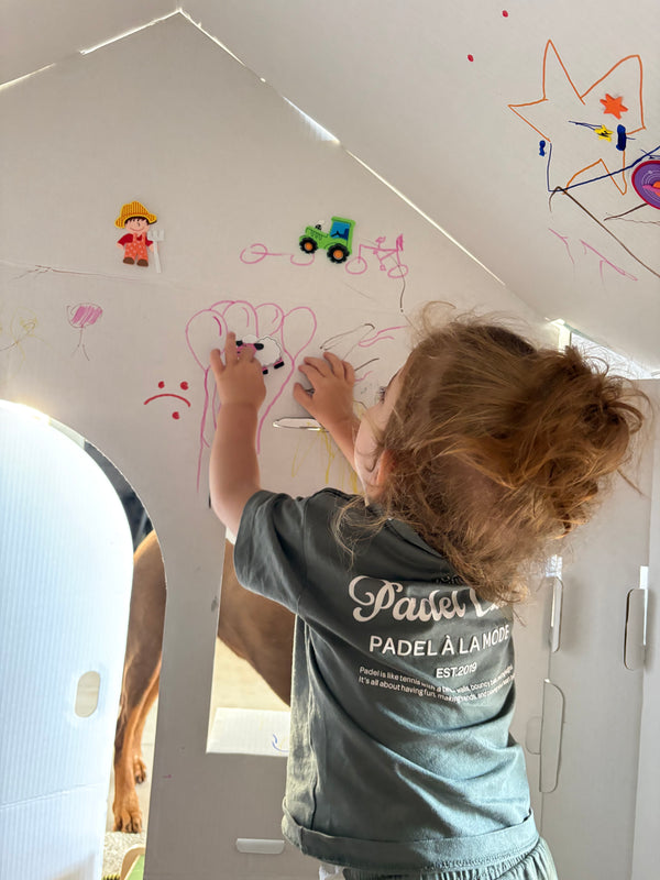 A young child with curly brown hair is seen from behind, wearing a gray t-shirt and drawing on a white wall with markers. 