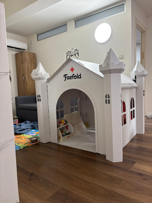 A white castle-shaped children's playhouse by Faefold, set up in a home interior with wood flooring and a dark sofa in the background.