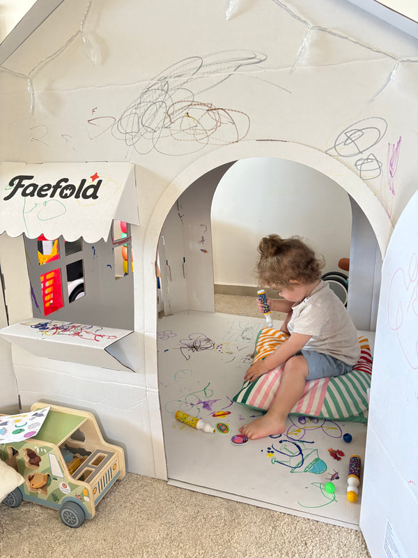 A child sits inside a white house-shaped play tent with an archway, colorful drawings scribbled on the walls, and a small counter with a Faefold sign on the left. The child is on a striped cushion on the floor, with toys scattered around in a playroom setting.
